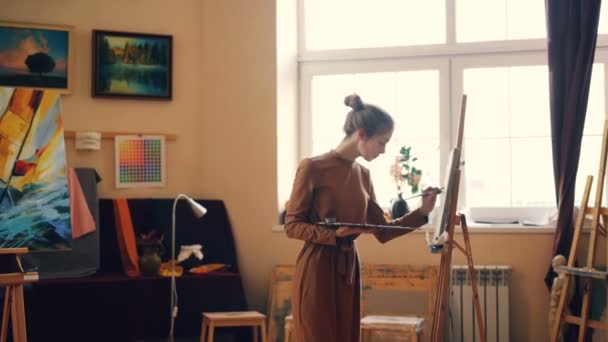 Die hübsche junge Malerin arbeitet im Atelier mit Palette und Malerei auf Leinwand, die sich auf kreative Arbeit konzentriert. Einsamkeit, Kreativität und Talentkonzept. — Stockvideo
