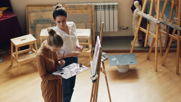 在现代艺术工作室里, 两个年轻的女艺术老师和学生在调色板上混合颜色的高角度的视角, 说话和微笑, 然后在画布上作画. — 图库视频影像