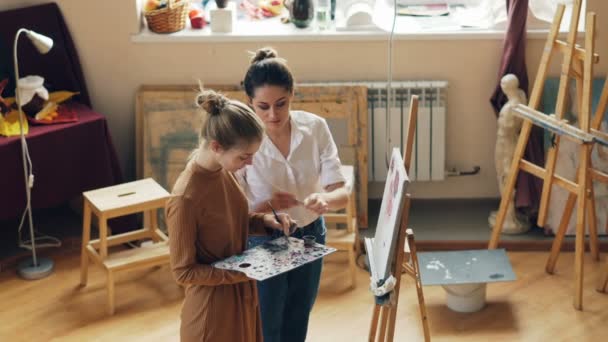 Good-looking young woman is holding palette and painting flowers under guidance of helpful teacher standing together in workroom of art school and talking. — Stock Video