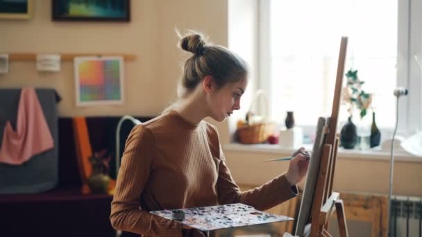 Ładna Blondynka jest malowanie piękne kwiaty na płótnie pracy w studio sam, stojąc przed sztalugi z pędzlem i palety i jasne obrazy przedstawiające. — Wideo stockowe