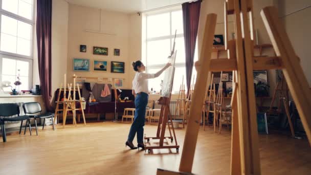 Young female painter is painting picture indoors working inside light workshop alone. Wooder easels, authentic artworks and arts tools are visible. — Stock Video