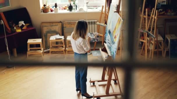 La joven creativa está ocupada pintando la imagen trabajando sola en un estudio moderno con muebles de madera, caballetes y herramientas de arte. Creación de un concepto de obra maestra . — Vídeos de Stock