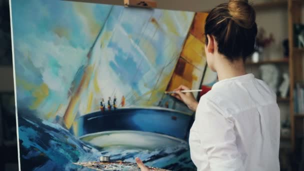 Skilled artist pretty woman is painting seascape sea waves and boat with acrylic paints on canvas holding palette and brush creating masterpiece. People and arts concept. — Stock Video