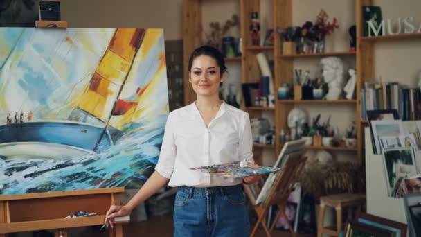 Retrato de una pintora confiada sosteniendo paleta y pincel de pie cerca del paisaje marino mirando a la cámara y sonriendo. Taller moderno con obras de arte está en el fondo . — Vídeo de stock