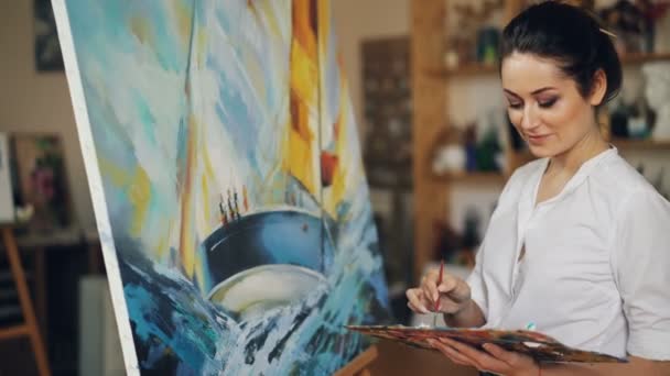 Mujer joven sonriente está pintando cuadro que representa el barco y las olas azules del mar sobre lienzo en pinturas al óleo, ella está sosteniendo el pincel y la paleta de pie cerca del caballete . — Vídeos de Stock