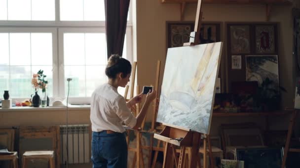 Orgullosa artista está tomando una foto de su obra maestra utilizando la pantalla táctil de la cámara del teléfono inteligente y mirando el dispositivo de pie en el taller. Concepto de fotografía y artes . — Vídeo de stock