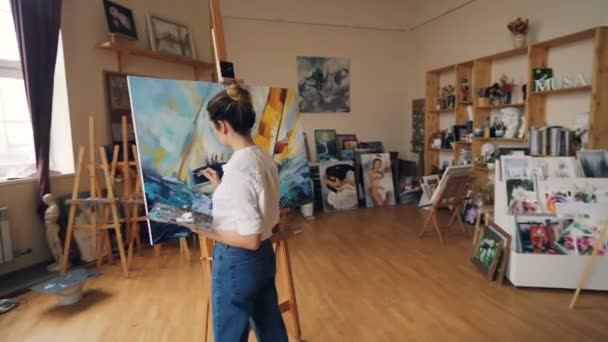 Pan shot of serious girl professional painter working in studio painting marine landscape with tempera paints holding palette and brush. Artworks and creativity concept. — Stock Video