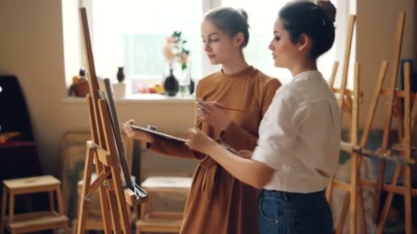 Professor profissional de arte escola está trabalhando com diligente menina estudante pintura quadro e falando partilha experiência dentro de casa na sala de trabalho cheia de obras de arte . — Vídeo de Stock