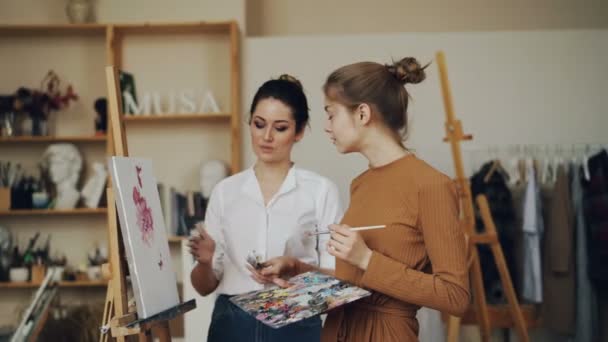 Alegre joven profesora y estudiante están discutiendo obras de arte juntos hablando y mirando la imagen. Taller moderno con caballetes y herramientas es visible . — Vídeos de Stock