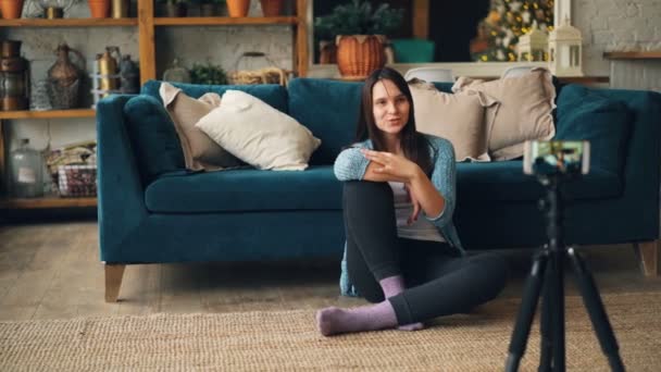 Emocjonalne kobieta młody blogger jest nagrywanie wideo dla jej vlog, rozmawiając i patrząc na aparat w smartfonie. Dziewczyna siedzi na podłodze w domu noszenia odzieży casual. — Wideo stockowe