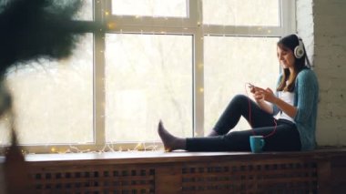 Mutlu genç kadın evde pencere pervazına üzerinde oturan müzik dinleme akıllı telefon ve kulaklık kullanarak. Modern teknoloji, millennials ve Gençlik Kültür kavramı.