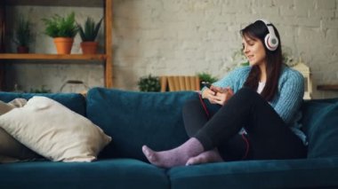 Genç hanımefendi smartphone kullanarak kulaklık üzerinden müzik dinlediği, dinlenme ve konfor zevk gülümseyen ve gülüyor kanepede oturan ev.