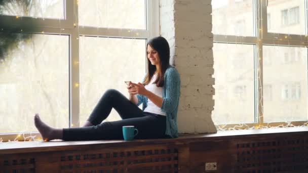 Cheerful young woman is using smartphone sitting on window sill with cup of tea relaxing at home and enjoying modern technology. People and gadgets concept. — Stock Video