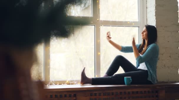 Vrolijke jonge dame is het maken van online video-oproep zittend op de vensterbank kijken naar smartphone scherm glimlachend zwaaien hand en praten. Communicatie en technologie concept. — Stockvideo