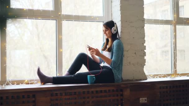 Brune fille mignonne est assis sur le rebord de la fenêtre, écouter de la musique dans les écouteurs et en utilisant un smartphone. Intérieurs modernes, technologie et concept de personnes relaxantes . — Video