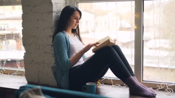 Krásná dívka čte knihu obracet stránky sedí na okně siil a někdy při pohledu z okna. Literatura, koncept mládeže kultury a interiéry. — Stock video