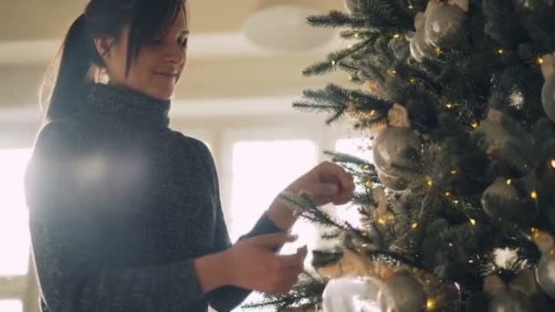 La giovane donna felice sta decorando l'albero di Capodanno preparandosi per il giorno di Natale a casa creando un'atmosfera festiva. Sfere e luci lucenti sui rami sono visibili . — Video Stock