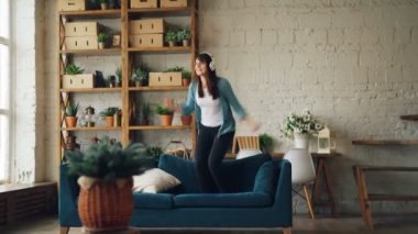 Komik kadın dans ve eğlence ve zevk özgürlük ve boş zaman sahip kulaklık üzerinden müzik kanepe üzerinde atlama. Loft tarzı daire arka planda olduğunu.