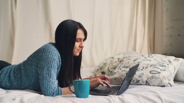 Leende dam ljuger i sängen hemma arbetar med laptop att skriva och tittar på skärmen, surfa på Internet. Modern teknik, människor och koncept. — Stockvideo