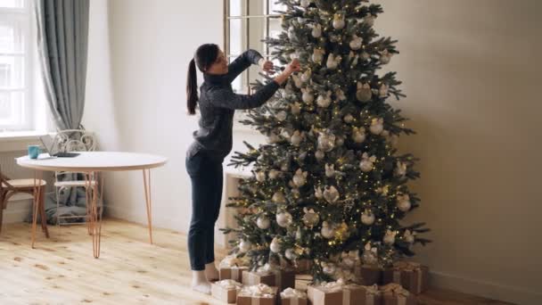 La giovane donna sorridente è impegnata a decorare l'abete per le vacanze di Capodanno toccando palle, giocattoli, luci e decorazioni creando un bel design. Ragazza indossa maglione e jeans . — Video Stock