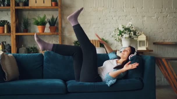 Snygg ung kvinna i casual kläder liggande på soffan, lyssnar på musik i hörlurar och dans rörliga ben och armar. Livsstil och ungdom koncept. — Stockvideo