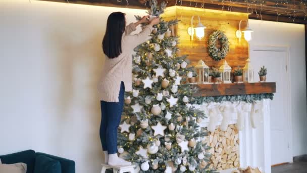 Slim ragazza in maglione e jeans sta decorando bellissimo albero di Capodanno toccando palle e stelle e creando un design perfetto. Caminetto decorato e luci sono visibili . — Video Stock