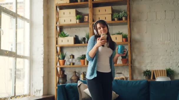 Glad tjej dansar hemma lyssnar på musik genom hörlurar och hålla smartphone tittar på skärmen. Underhållning, millennials och prylar koncept. — Stockvideo
