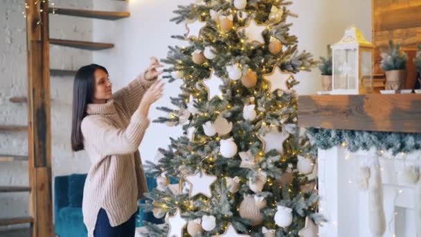 Chica sonriente está decorando el árbol de Navidad con bolas y luces disfrutando de las vacaciones y la actividad creativa. Hermosa habitación decorada con chimenea es visible . — Vídeos de Stock