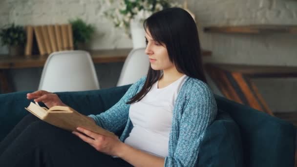 Menina calma está lendo livro sentado no sofá em casa virando páginas desfrutando de paz, literatura e conforto. Nice loft estilo quarto é visível no fundo . — Vídeo de Stock