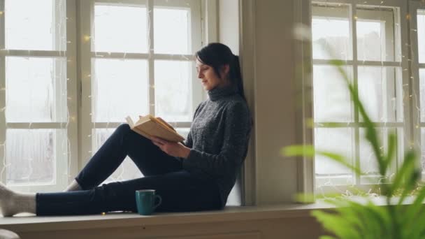 Bella bruna in maglione e jeans sta leggendo un libro seduto sul davanzale della finestra il giorno di Natale godendo di solitudine, tempo libero e vacanza. Concetto di letteratura e riposo . — Video Stock