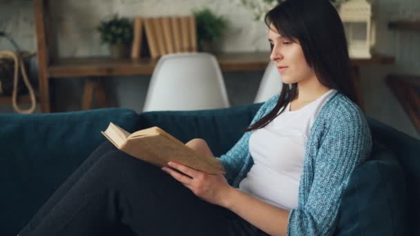 Jolie jeune femme lit le livre et sourit assis sur le canapé à la maison et se détendre seul. Littérature intéressante, hobby et concept de personnes intelligentes . — Video
