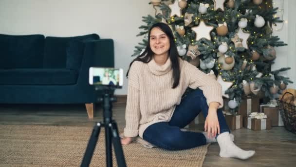 La giovane donna emotiva è seduta sul pavimento vicino all'albero di Natale e sta registrando video per il blog online utilizzando la fotocamera dello smartphone. Gril sta parlando e gesticolando . — Video Stock