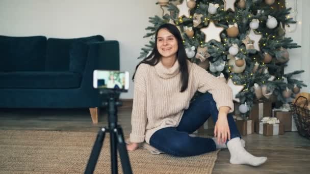 Emotionele jonge vrouw zit op de vloer in de buurt van de kerstboom en opnemen van video voor online blog met behulp van de camera van de smartphone. Gril is praten en gebaren. — Stockvideo