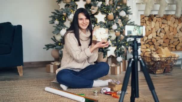 Femme gaie vlogger enregistre tutoriel sur l'emballage cadeau assis près de l'arbre de Noël et tenant papier d'emballage lumineux, rubans, boîtes et ciseaux . — Video
