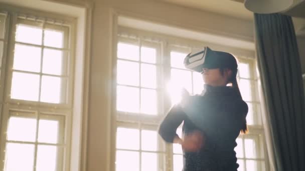 Smějící se mladá dívka v svetru se baví s virtuální realita brýle pohybující se ruce ve světelné studio s brýlemi na hlavě a užívat si miniaplikaci. — Stock video