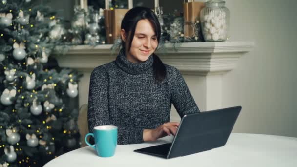 Jovem sorridente de camisola está trabalhando com laptop no Natal sentado perto da árvore de Ano Novo pensando e digitando. Conceito moderno de tecnologia e férias . — Vídeo de Stock