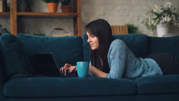 Mujer joven atractiva en ropa casual está escribiendo en el ordenador portátil y sonriendo mientras está acostado en el sofá en el estudio de estilo loft. Tecnología moderna y concepto de los millennials — Vídeos de Stock