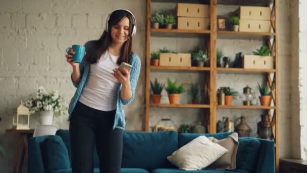 Ung brunett lyssnar på musik genom hörlurar och använda smartphone tittar på skärmen och leende att ha kul dans och vrida inomhus på hem. — Stockvideo