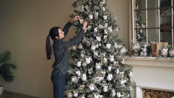 Smiling young woman is decorating green Christmas tree with beautiful balls creating authentic design getting ready for winter holidays. People and interior concept. — Stock Video