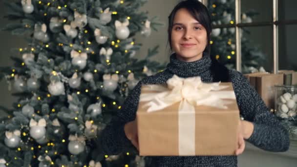 Portrait of beautiful brunette giving present on Christmas day standing indoors at home with decorated New Year tree and mantel in background. Gifts and people concept. — Stock Video