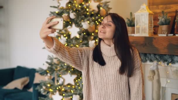 Jovem feliz está falando com amigos on-line fazendo videochamada com smartphone no dia de Natal de pé perto do abeto e decorado mantel em casa . — Vídeo de Stock