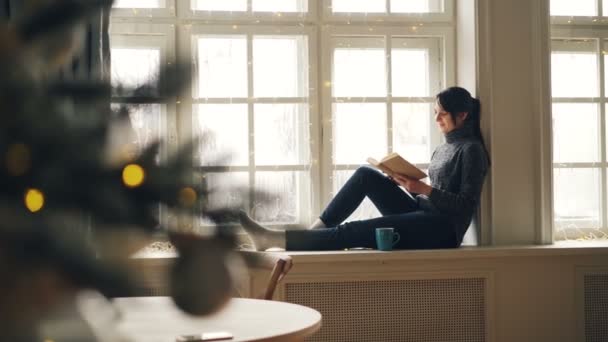 Улыбающаяся молодая женщина читает книгу, сидя на подоконнике в Рождество и наслаждаясь отдыхом и мирным отдыхом. Видны украшенные елки и огни . — стоковое видео