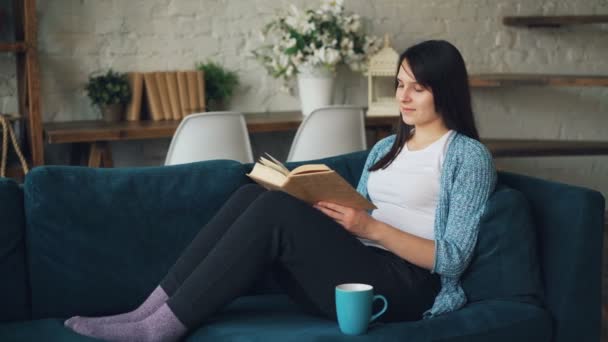 きれいな女性は、紅茶とリラックスの自宅のソファの上に座って近代文学を楽しんで本を読んでいます。家、新世紀教育コンセプト. — ストック動画