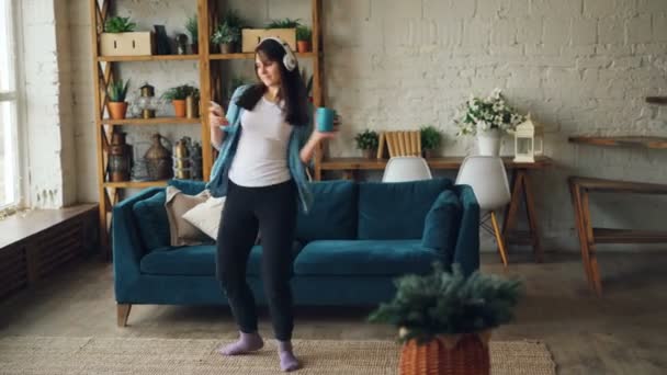 Aantrekkelijke brunette is plezier thuis luisteren naar muziek via de koptelefoon en dansen bedrijf smartphone en cup. Moderne technologie en mensen concept. — Stockvideo