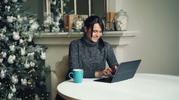 Chica feliz está trabajando con el ordenador portátil en el día de Navidad sentado en la mesa cerca del árbol de Año Nuevo decorado y escribiendo mirando la pantalla de envío de correos electrónicos a los amigos . — Vídeo de stock