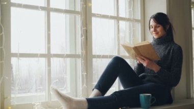 Rüya gibi genç kadın kitap ve Noel pencere-pervazına üzerinde oturan gülümseyen aydınlatma ile dekore edilmiş okuyor. Kız kazak ve trendy kot giyiyor.