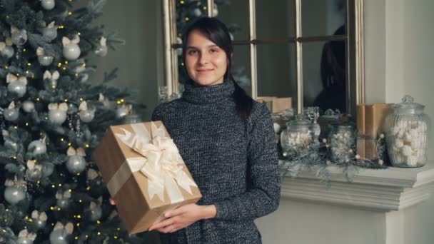 ギフト ボックスに立って魅力的な若い女性の肖像画付近のクリスマス ツリーと装飾のマントルピースとカメラを見て笑っています。新しい年の日とプレゼントのコンセプト. — ストック動画