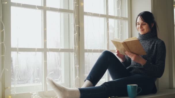 Giovane donna sognante sta leggendo il libro e sorridente sul Natale seduto sul davanzale decorato con illuminazione. Ragazza indossa maglione e jeans alla moda . — Video Stock