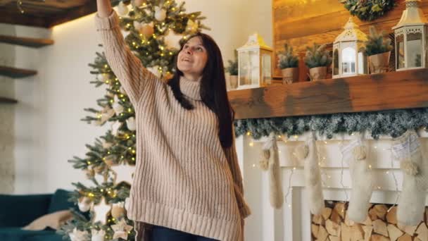 Chica despreocupada se está tomando selfie en el día de Navidad de pie cerca de árbol de Año Nuevo y chimenea sosteniendo el teléfono inteligente y posando para la cámara. Concepto de vacaciones y fotos . — Vídeo de stock