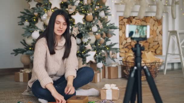Mujer bloguera hábil está enseñando a sus seguidores a decorar regalos de Navidad sentados en el suelo sosteniendo la caja presente y la cinta y grabando video con teléfono inteligente . — Vídeos de Stock